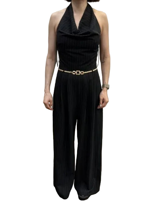 A woman modeling a black sleeveless halter jumpsuit with a cowl neckline, pinstriped wide-leg pants, and a thin gold chain belt with a central gold ring, standing in an indoor room with a concrete floor and off-white wall.